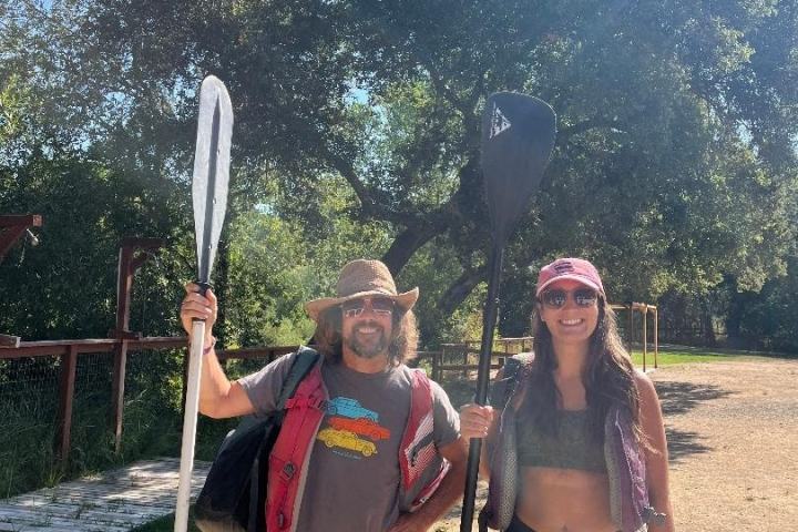Two people in outdoor gear holding paddles, standing on a dirt path with trees in the background.